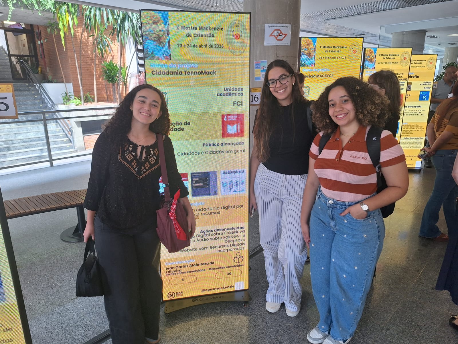 Três alunas participantes do Cidadania TecnoMack diante do painel digital do projeto na X Mostra Mackenzie de Extensão.